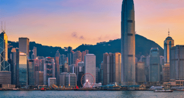 Hong Kong skyline at sunset, featuring skyscrapers and a waterfront with a Ferris wheel, set against vibrant orange and pink skies.