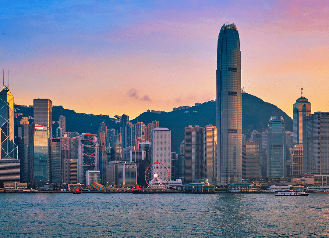 Hong Kong skyline at sunset, featuring skyscrapers and a waterfront with a Ferris wheel, set against vibrant orange and pink skies.