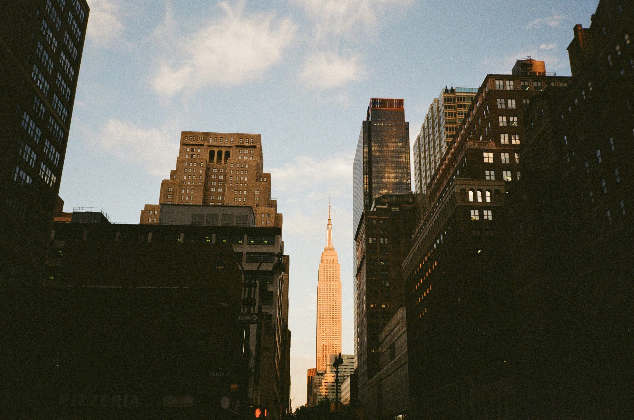 City street view at sunset with tall buildings on both sides and a prominent skyscraper in the background, bathed in warm light.